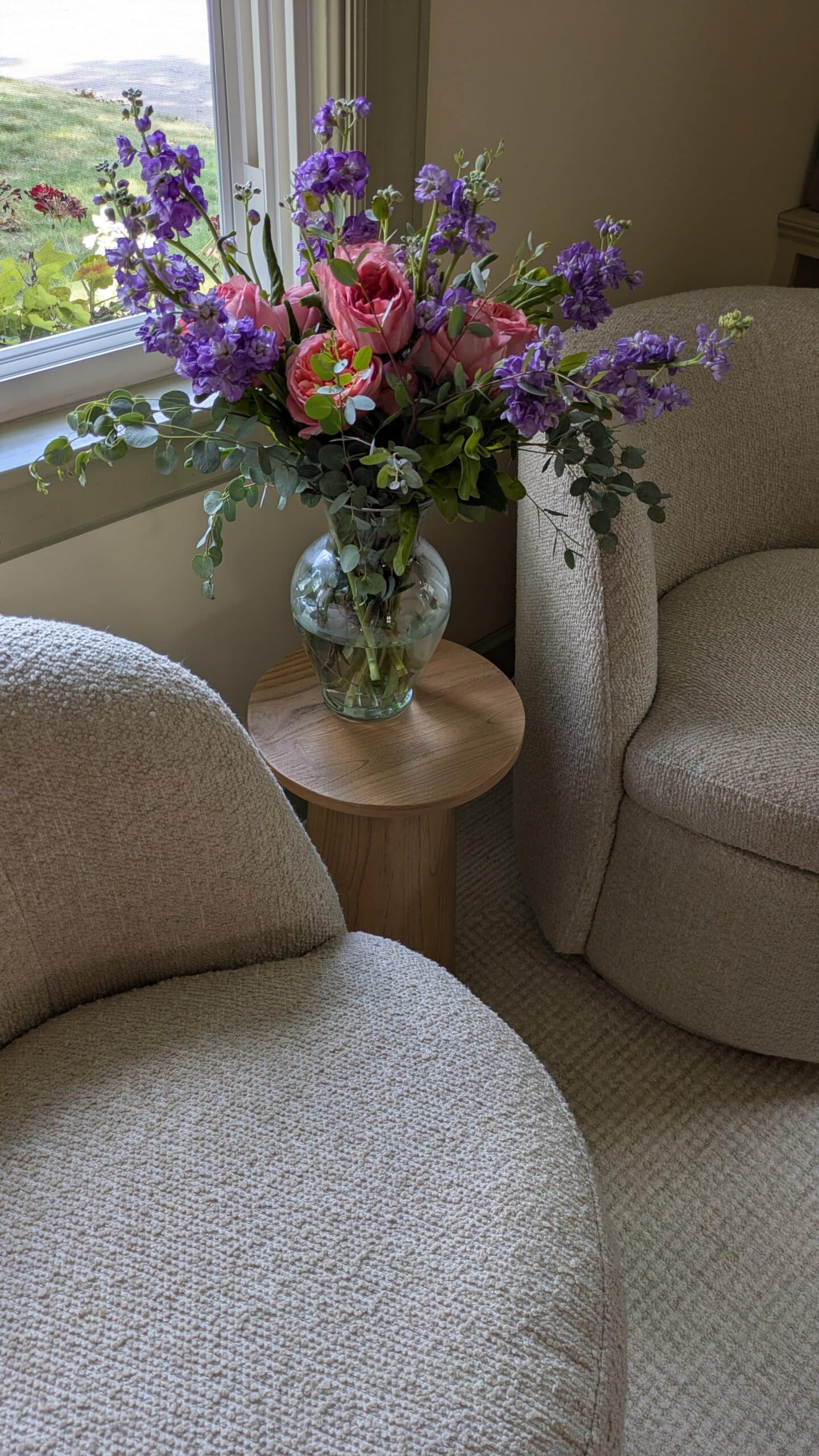 Two accent chairs near a window with a accent table in the middle and a vase with beautiful flowers.