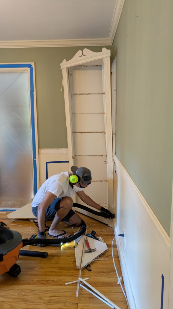A man working with a shop vacuum renovating a dining room with a DIY dust barrier zipper cover in the background. He is removing a built in from the wall.