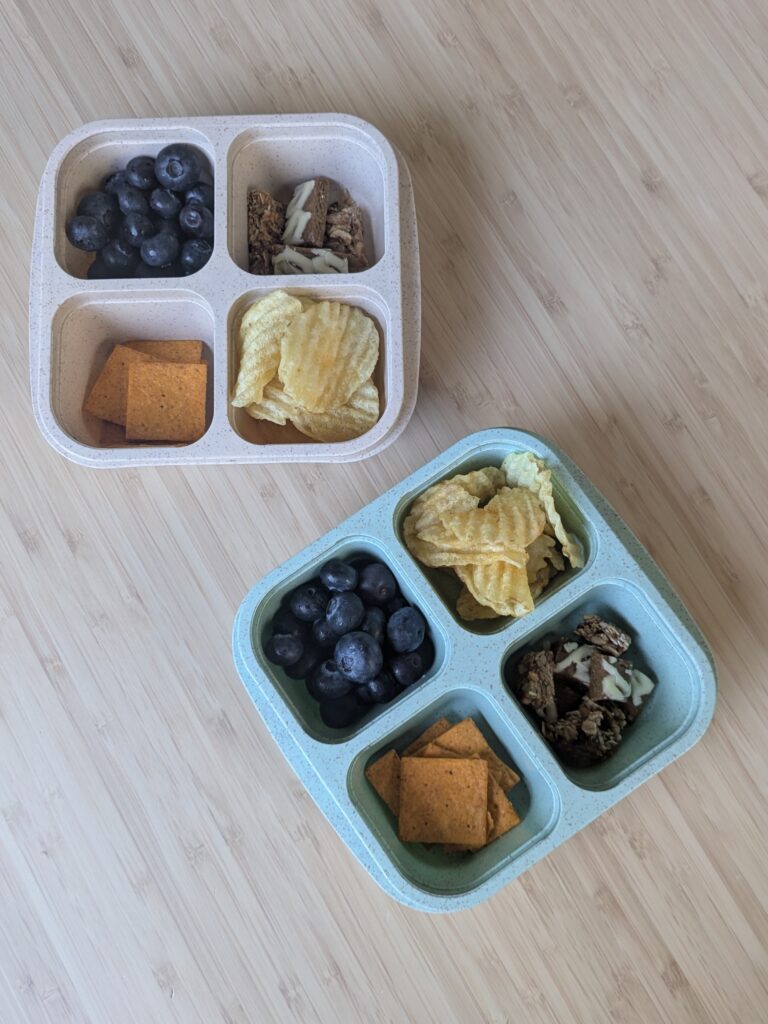 Four compartment kids snack container holding snacks such as blueberries, granola, crackers and potato chips.