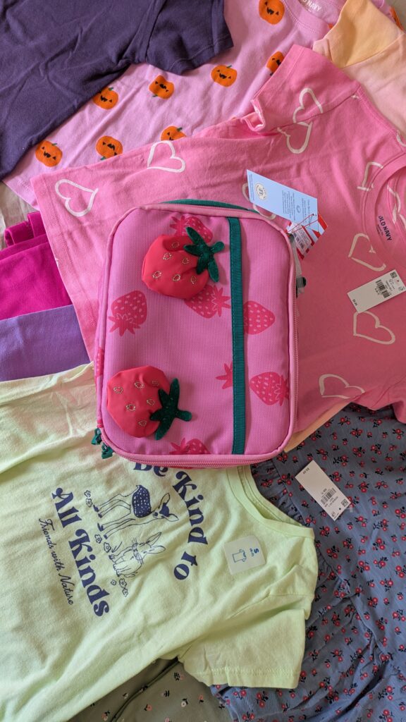 Fun prints and patterns of cotton clothing for girls laid out on a bed. Including a strawberry lunch bag.