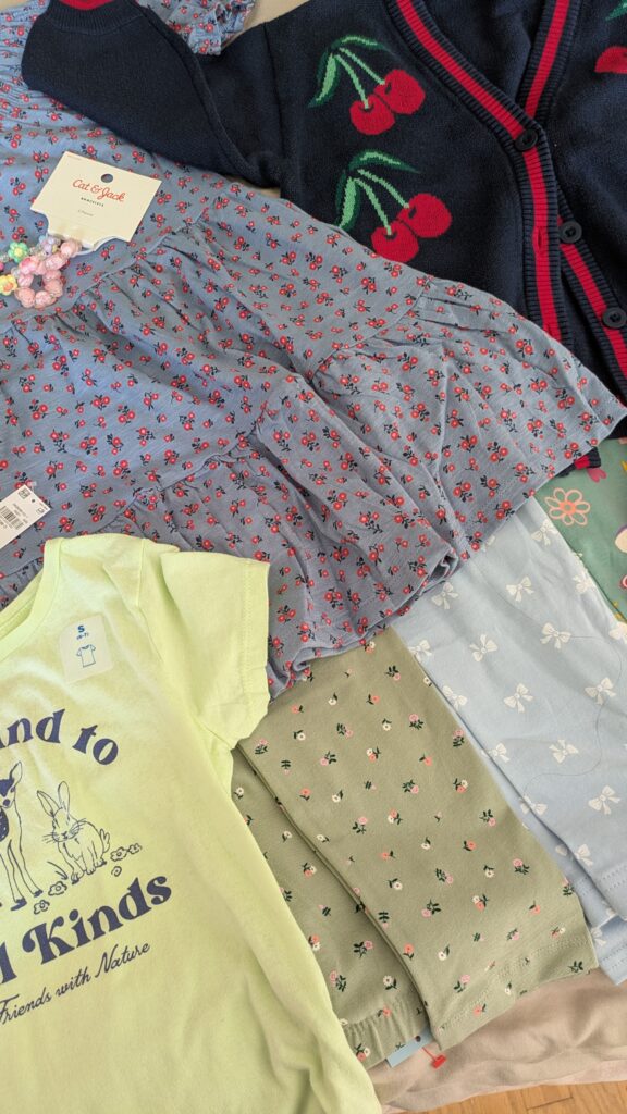 Fun cotton clothing for girls laid out on the bed including a dress and a cherry print cardigan as well as cute beaded bracelets from Target and Old Navy.