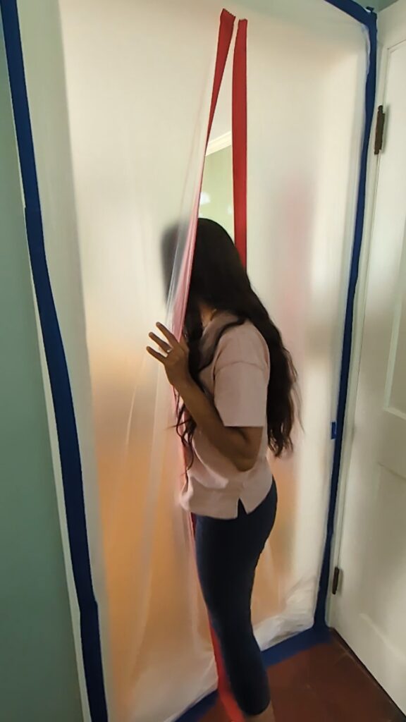 A plastic sheet on a door with a red zipper going down the middle. A woman going through the DIY door dust barrier zipper.