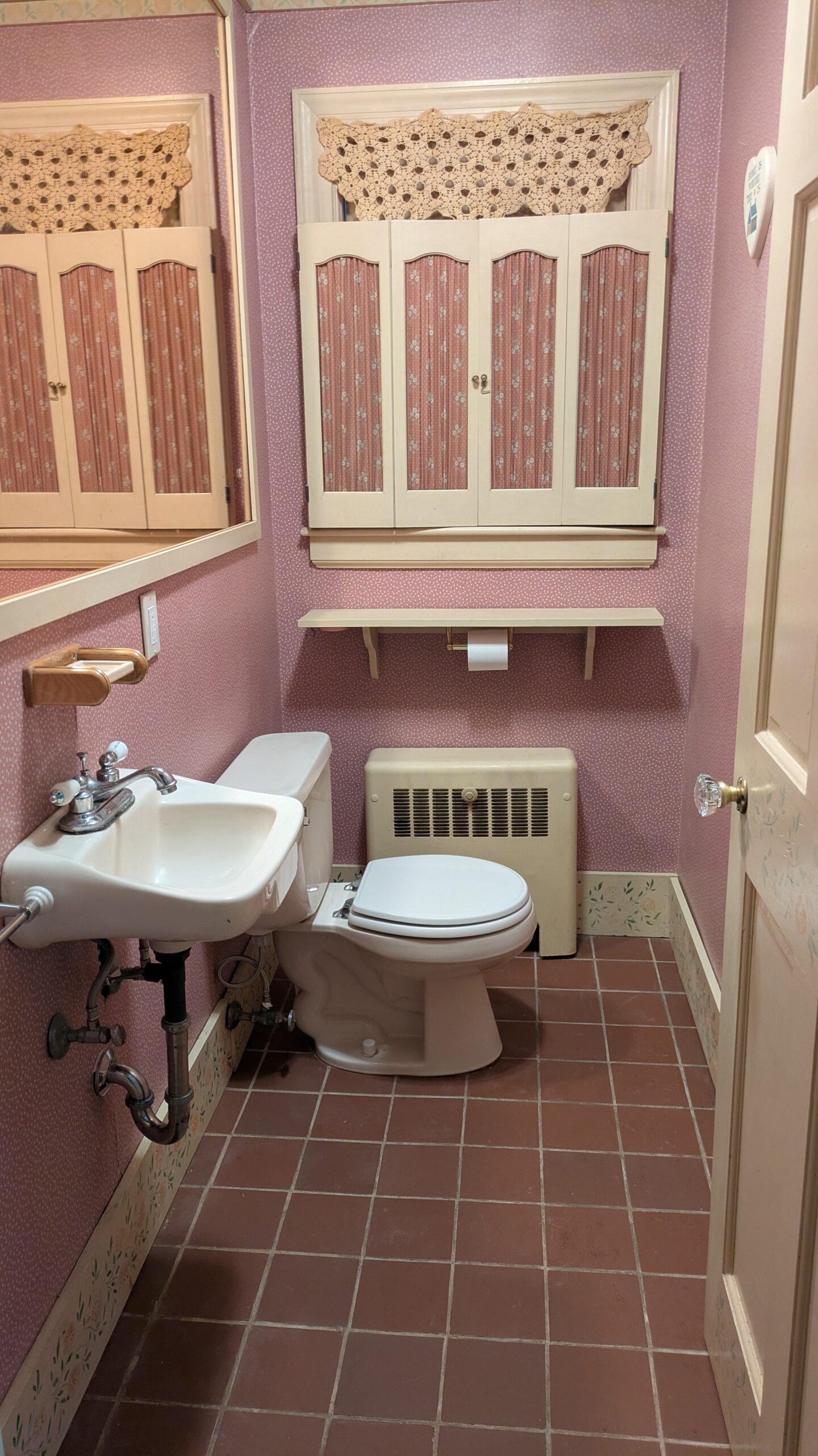 Dated pink half bathroom with pink wallpaper, pink shutters and large mirror, tiny sink and old fixtures.