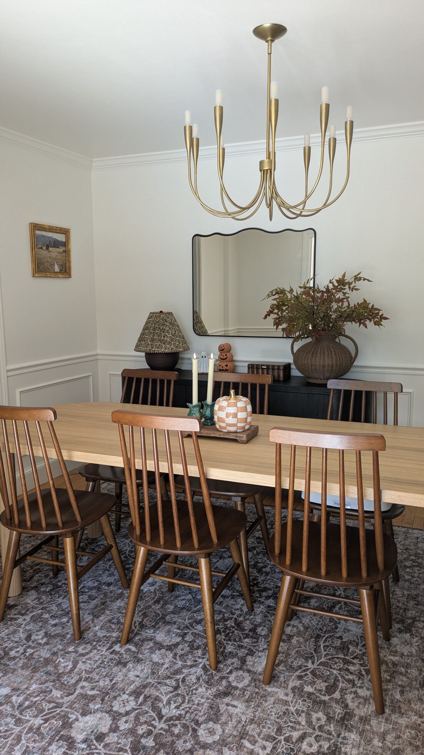 View of a dining room. With a black sideboard. A brown lamp base and a floral lamp shade on the sideboard. Fall and Halloween decor and faux Fall folliage as well as wall art in the room. Also shown three walnut spindle back chairs and an oak dining table.