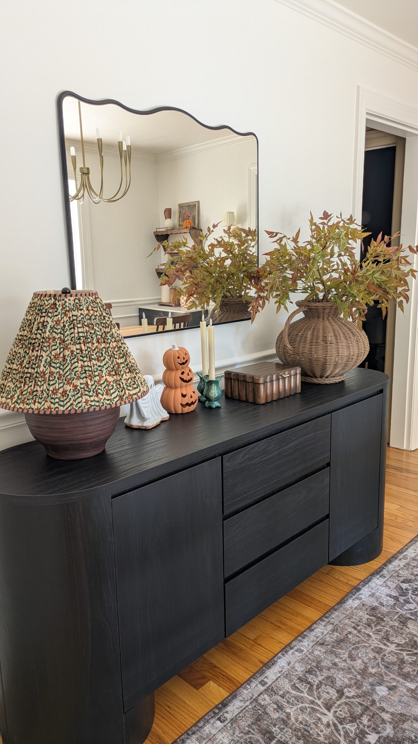 Black sideboard large wavy mirror. Large cottage floral print lamp and Fall / Halloween decor. Brown scallop wooden box and a large wicker vase with faux Fall stems.