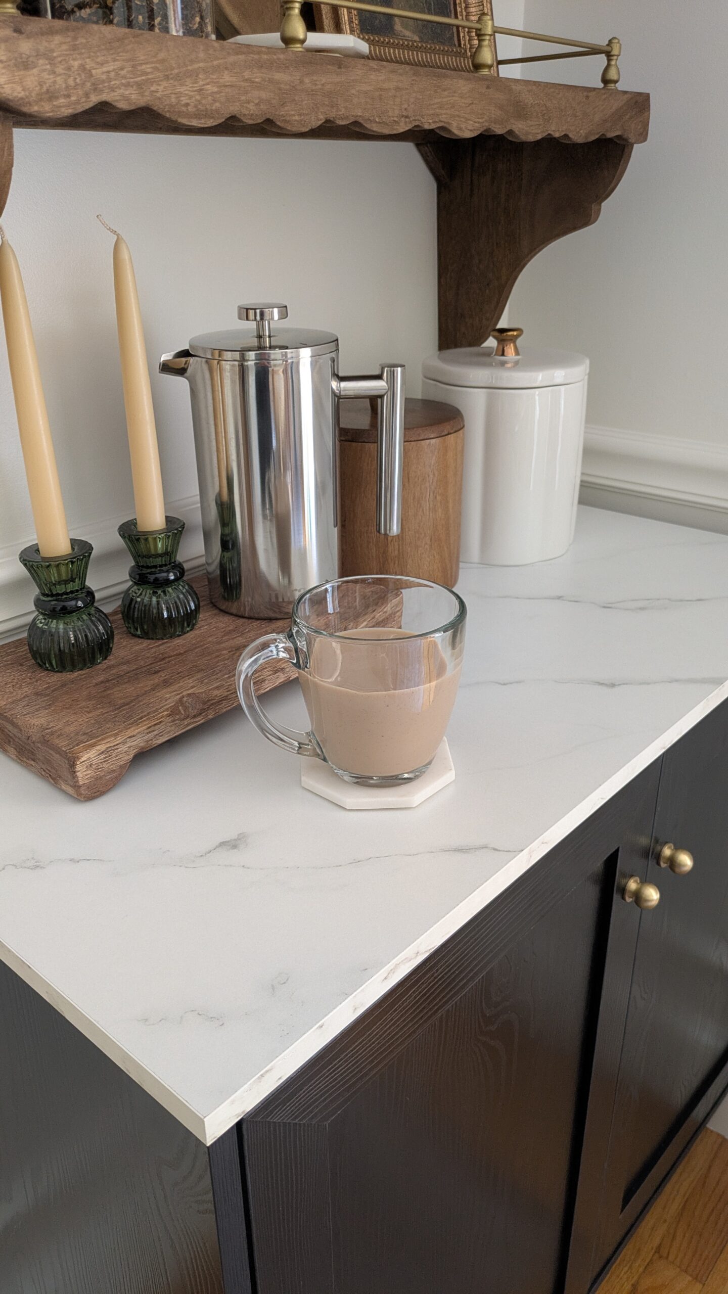 A cup of coffee in a glass mug, next to two jars one wooden and one ceramic also two candles in green glass votive and a stainless steel french press on a wooden riser. 