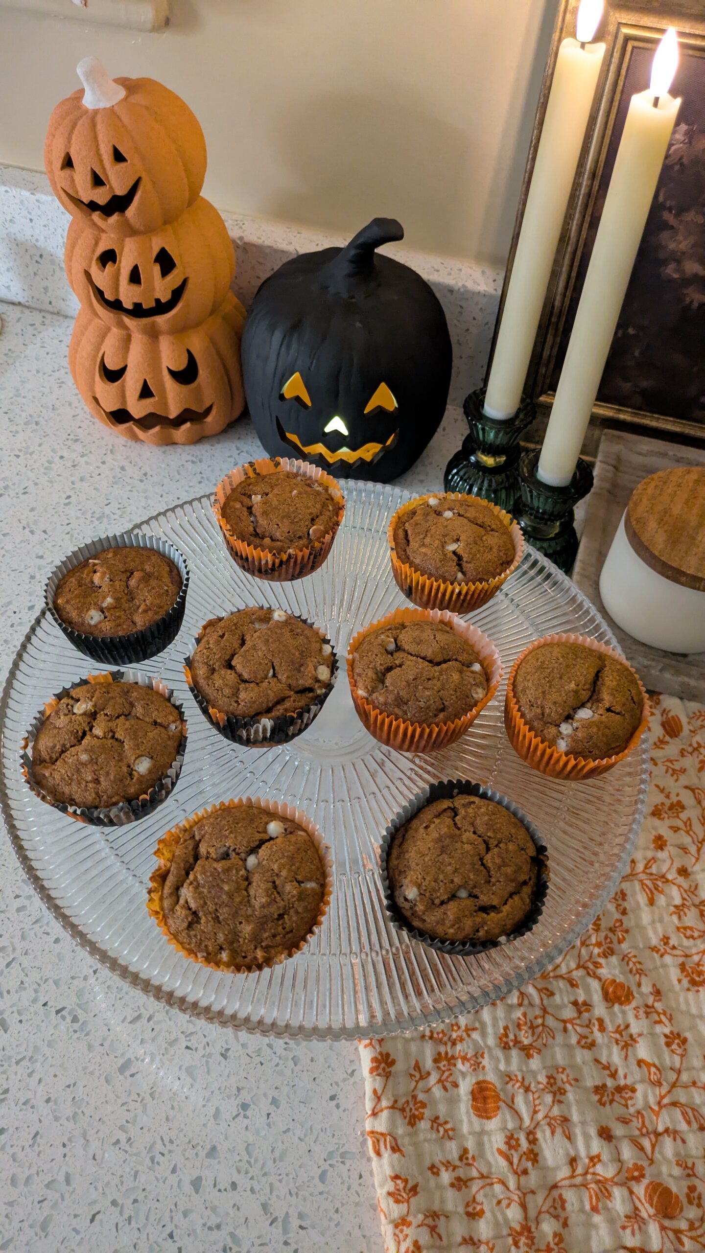 Pumpkin muffins on a glass cake stand. With stacked pumpkins, jack o lantern and candles decor on a counter top.