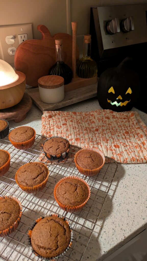 Muffins made with Simple Mills Pumpkin Muffin Mix on the counter resting on a cookie sheet. Fall cream colored towel with orange pumpkins and leaf art and glowing lit jack o lantern. salt cellar, oil and vinegar holder on a marble tray and a essential oil diffuser on the side.