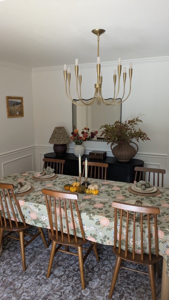 Hosting essentials - image of a dining room with a modern chandlier on the ceiling. a pasture wall art to the left wall. A wavy mirror on top of a sideboard and a cute floral pattern lamp and fresh dahlias in a vase on the sideboard as well as a scalloped wooden box and a large rattan vase with fauxe Fall stems. The dining table has a green floral pattern table cloth and brown woven chargers with organic off white dinner plates and floral napkins. There is a pumpkin and candles center piece in the middle.