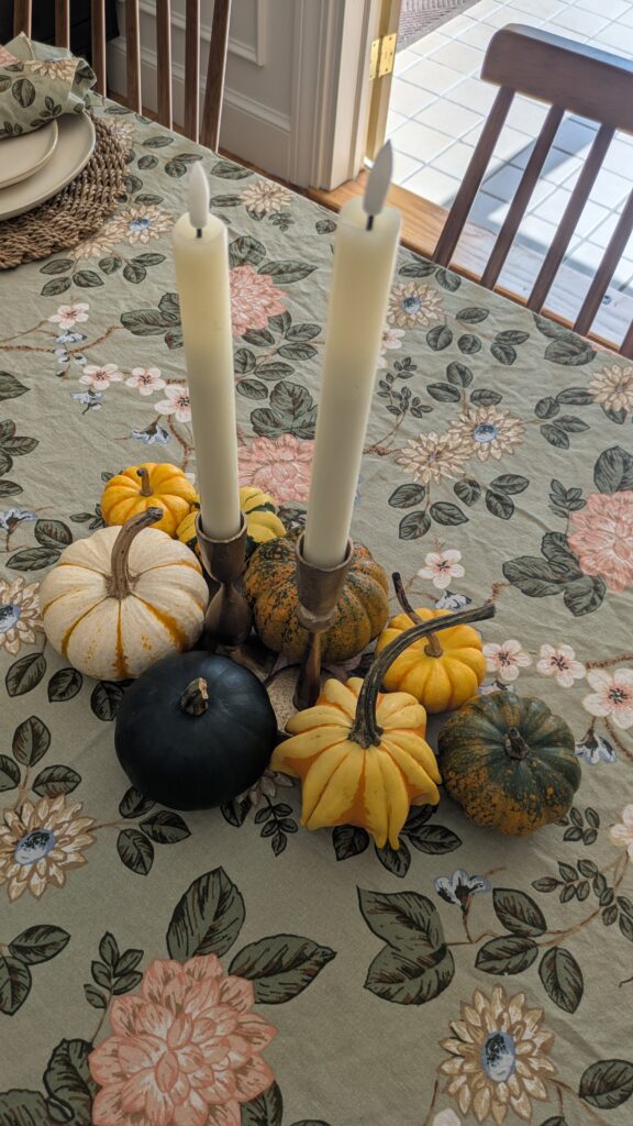 Hosting essentials - Green floral table cloth with various small different size and color pumpkin as the center piece with two antique brass candle holders in the middle and two ivory colored flameless tapered candles.
