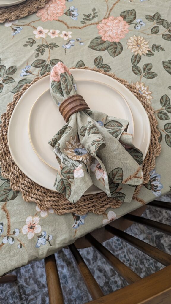 Hosting essentials - Floral green table cloth, rustic brown charger and organic off white dinner plate and salad plate on top with a floral napkin in a wooden napkin ring on a dining table.