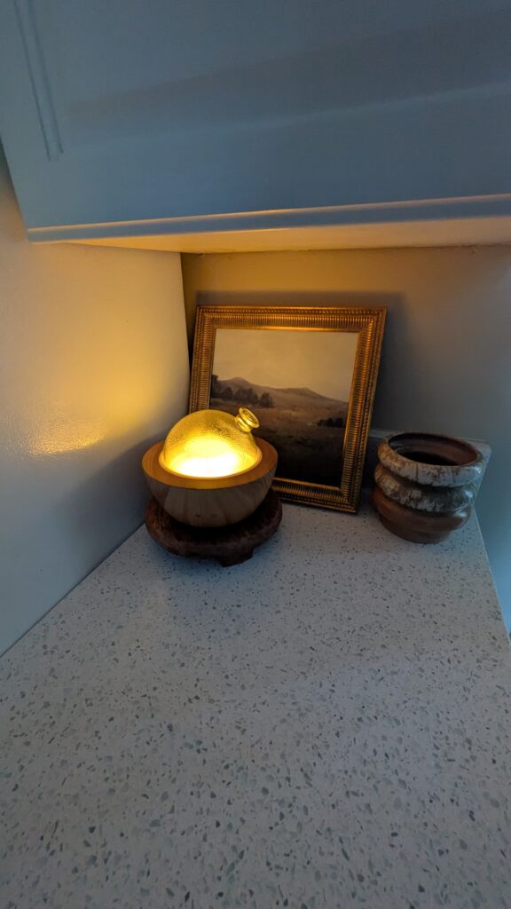 A diffuser glowing on the countertop. A picture of Fall pasture leaning against the wall with a ceramic brown and cream rustin pot next to the art.