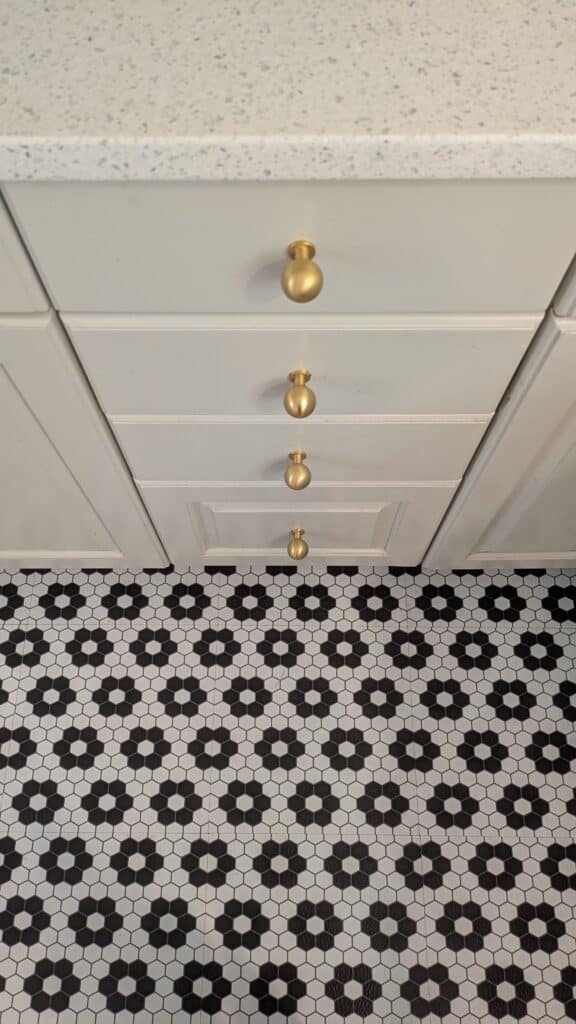 Goldknob on white cabinet drawers with a black and white floral tile on the ground.