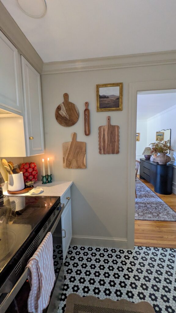 image of a corner of a kitchen also you can see the black sideboard and mirror and rug as well as lamp and a vase in the dining room. The kitchen was has serving boards hung up. On the counter there is a red tomato vase, wooden utensils in a utensilholder on top of a walnut lazy susan, a scallop shaped spoon rest and two green cabbage taper candle holders with flameless taper candles.