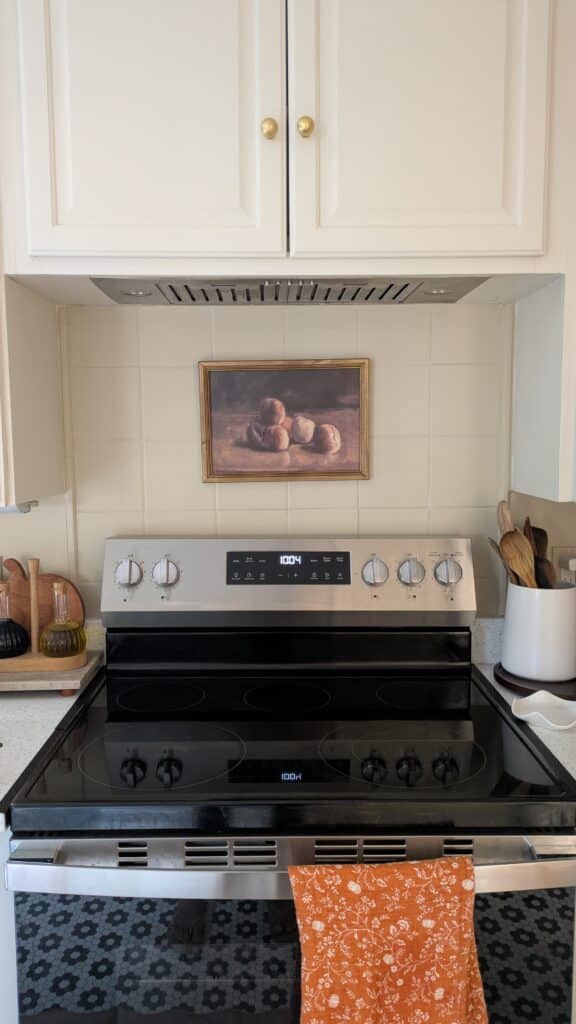 image of an electric stove, oil and vinegar holder on the countertop, utentsils in ceramic holder. Still life harvest art above the stove.