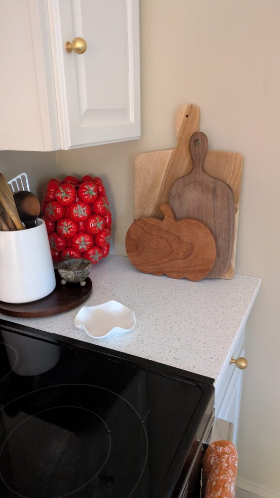 image of a red tomato vase, wooden utensils in a ceramic holder on a lazy susan, marble footed small bowl, scallop spoon rest on the counter and various size serving boards including a pumpkin shape one.