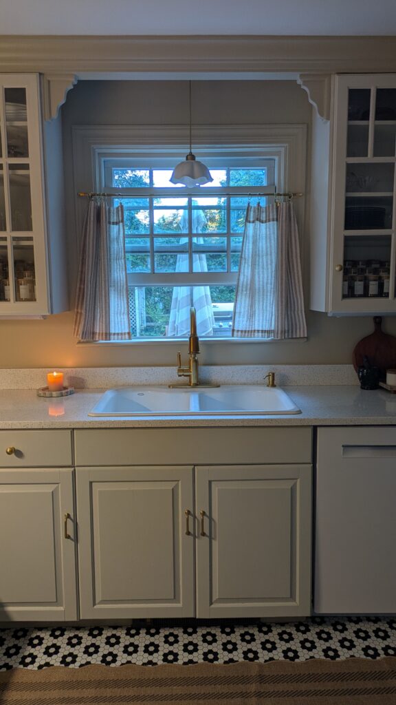 image of a kohler porcelain sink and cafe curtains on the window with a danty scallop white pendant light. Candle burning on the counterrop. Glass cabinetry showing spice jars. black and white floral pattern floor tiles with a scallop brown rug.