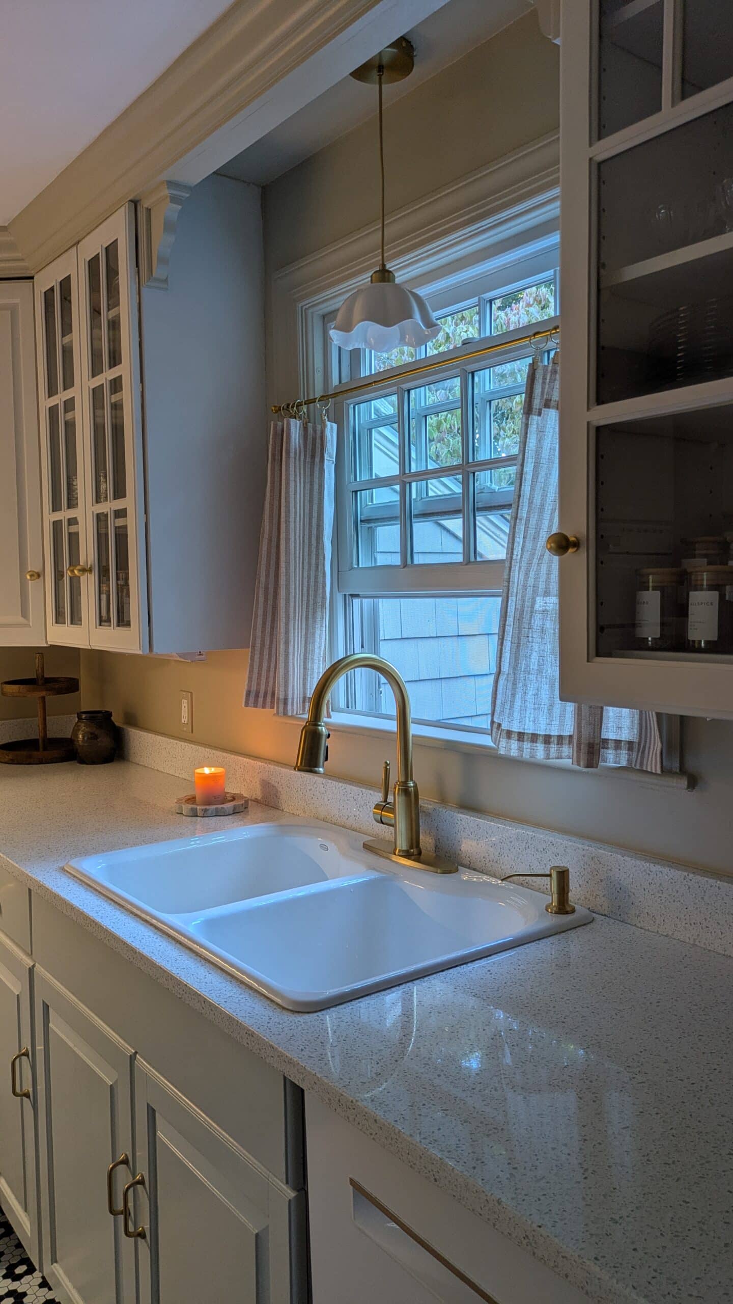 Beautiful brass fauct in a kohler porcelian sink. Cafe curtains on the kitchen window and glass cabinetry.