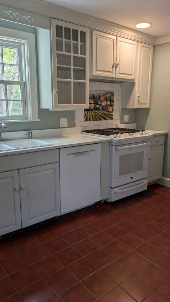 White kitchen a bit dated. Red old tiles on the floor. Tile mural above the stove.