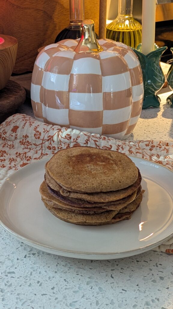 a stack of Simple Mills pumpkin pancakes on a cream color plate with a Fall towel tucked under and a checkered pumpkin in the background