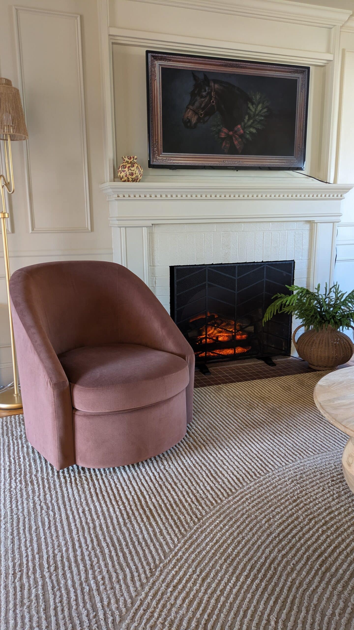 Accent chair in front of a fireplace. in the living room. Small decorative vase on the mantel. There is an image of a horse TV art over the mantel.