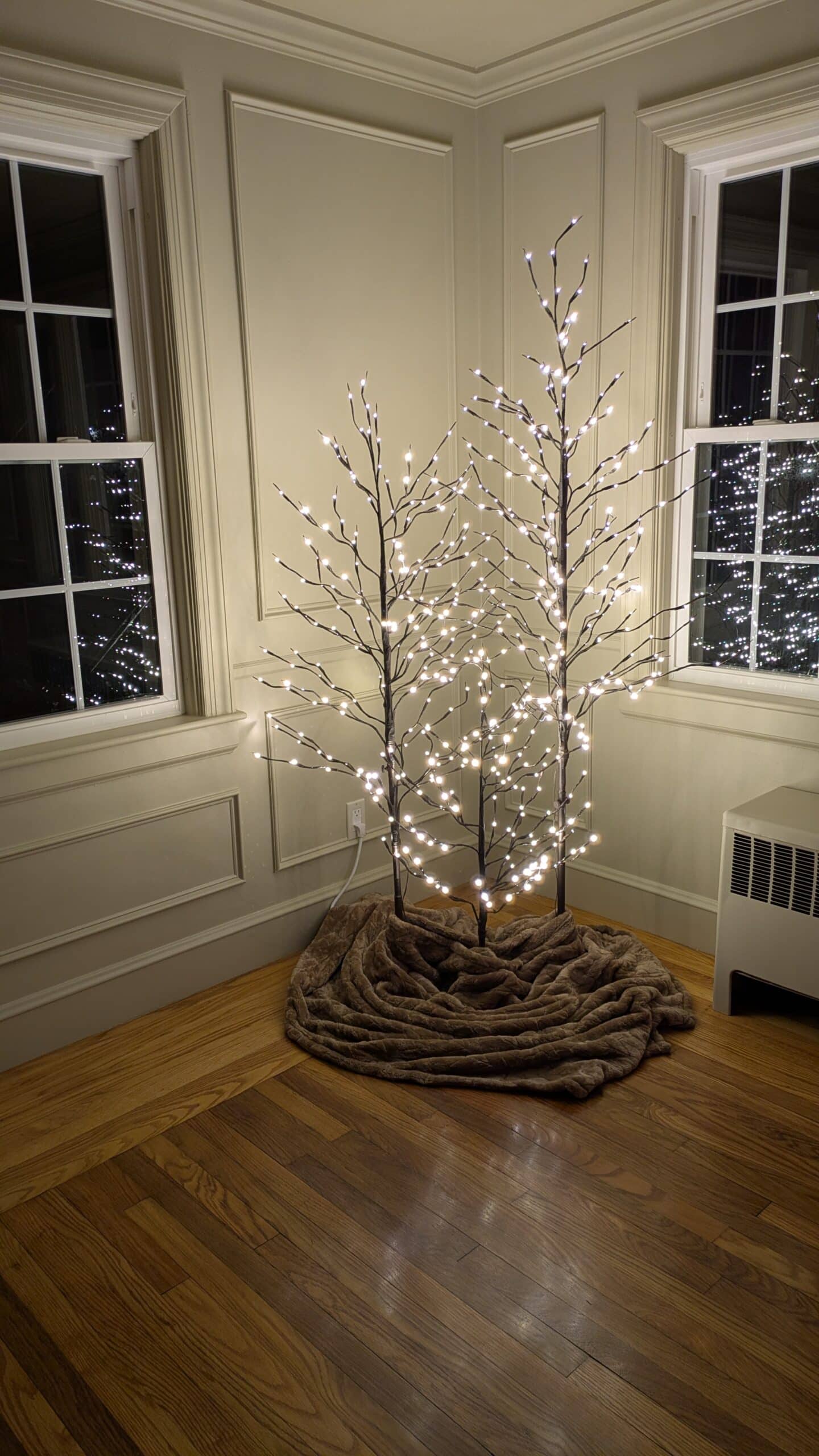 A set of three lighting trees in the living room. With a cozy blanket wrapped around it on the ground.